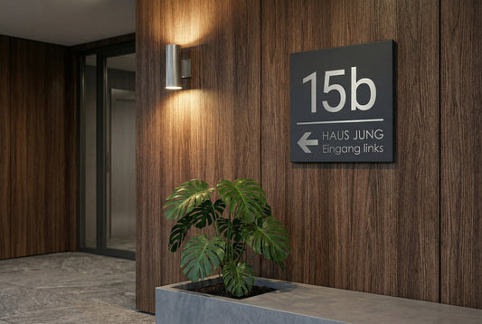 Anthrazit pulverbeschichtetes V2A Edelstahlschild mit Hausnummer 15b, Pfeil und Text „Haus Jung – Eingang links“ als moderner Wegweiser an dunkler Holzfassade im Eingangsbereich mit Pflanzkübel und Außenlampe