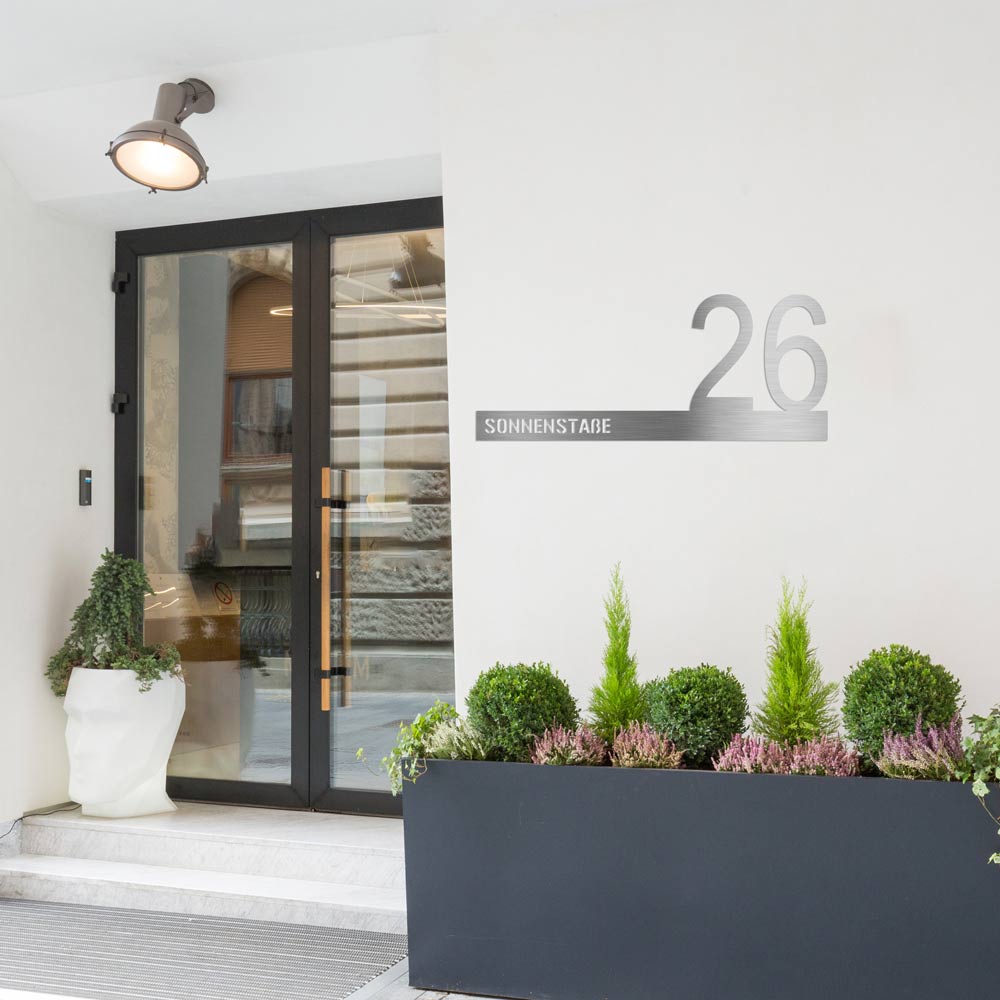 Personalisierbare Edelstahl-Hausnummer „Sonnenstraße 26“ an moderner Hausfassade mit Glastür und Pflanzkübeln. Minimalistisches Design, individuell anpassbar mit Wunschstraße und Hausnummer – ideal für stilvolle Eingangsbereiche.
