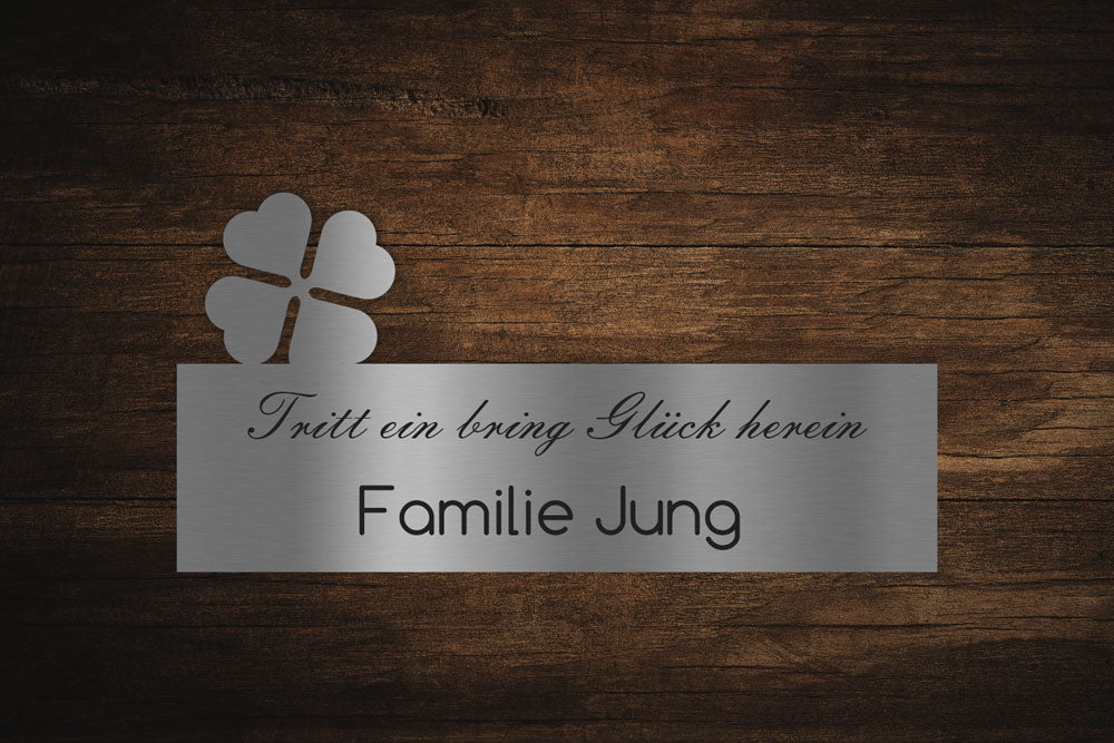 Metallschild mit Kleeblatt und Aufschrift „Tritt ein, bring Glück herein. Familie Jung“ auf dunklem Holzgrund.